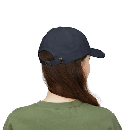 Back view of a person with long brown hair wearing a navy blue cap from the Worldwide PRIDE store, featuring a rainbow design and the embroidered pronouns she/her, along with an olive green sweatshirt. 
