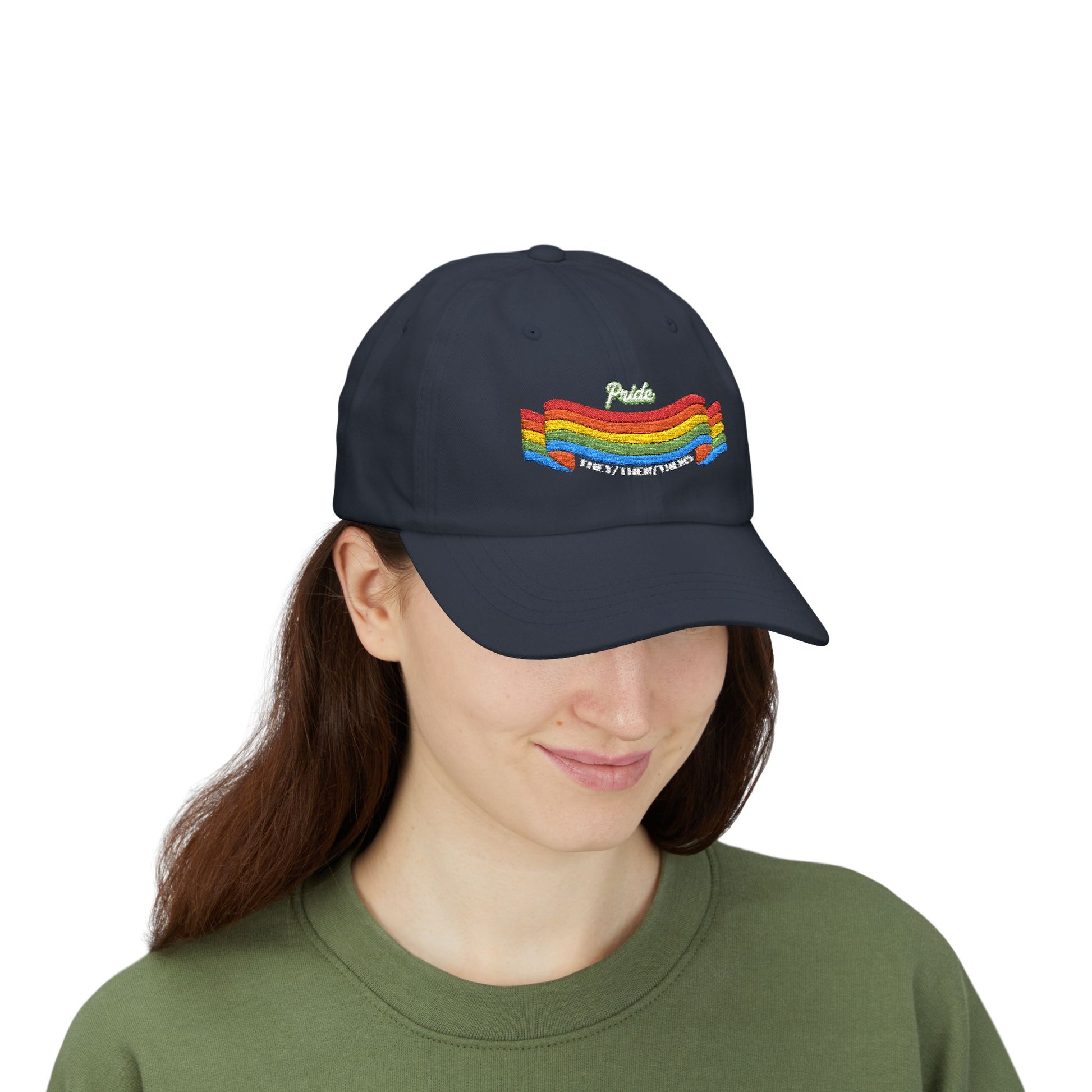 A person with long brown hair wears a navy blue cap from the Worldwide PRIDE store, featuring a rainbow design and the embroidered pronouns they/them/theirs, along with an olive green sweatshirt. The person is smiling softly and looking down.