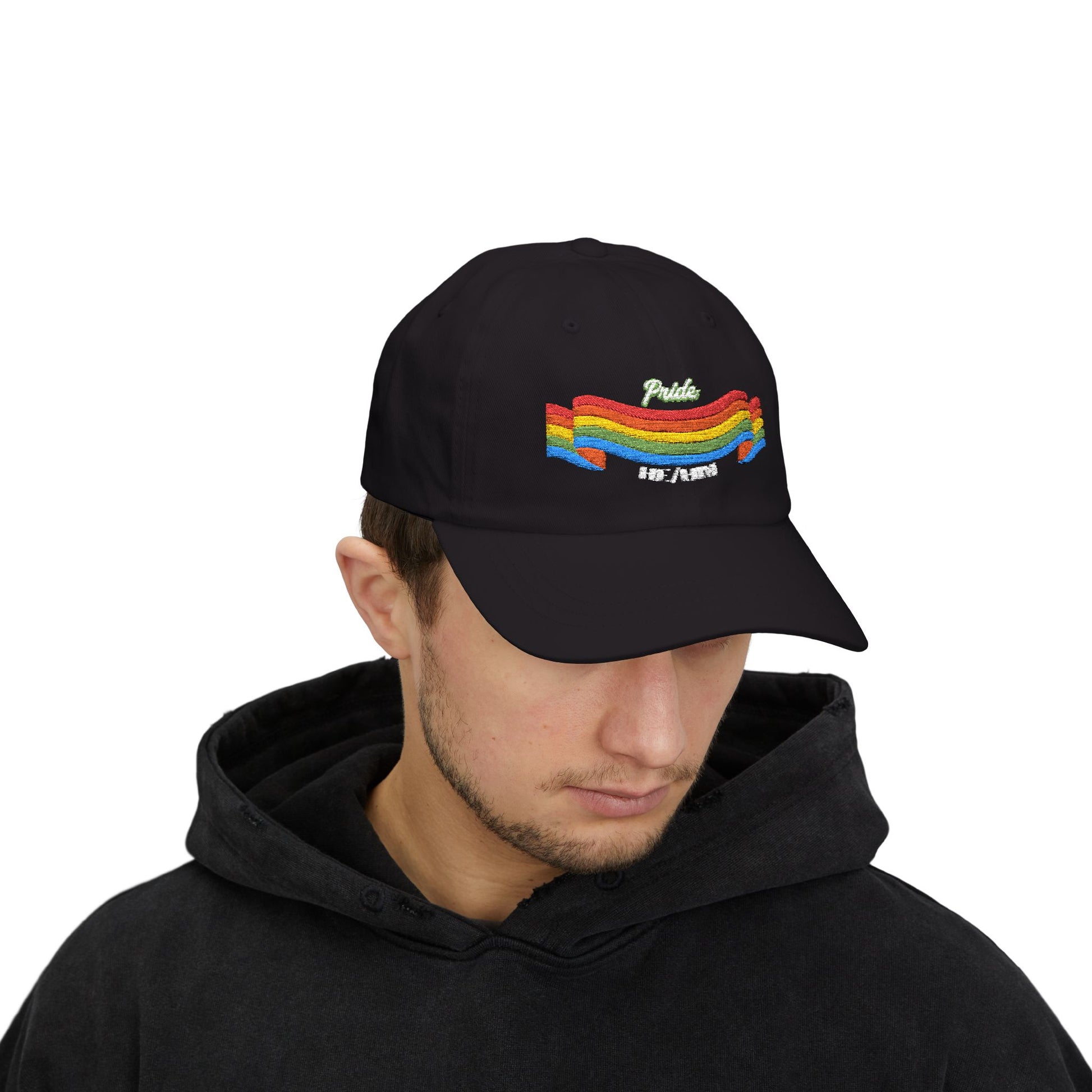 A person with short brown hair wears a black cap from the Worldwide PRIDE store, featuring a rainbow design and the embroidered pronouns he/him. The person is also wearing a black hooded sweatshirt and looking down.