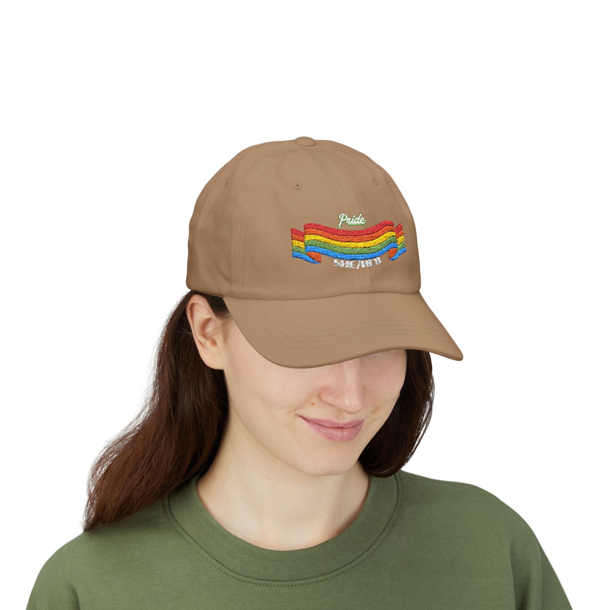 A person with long brown hair wears a khaki cap from the Worldwide PRIDE store, featuring a rainbow design and the embroidered pronouns she/her, along with an olive green sweatshirt. The person is smiling softly and looking down.