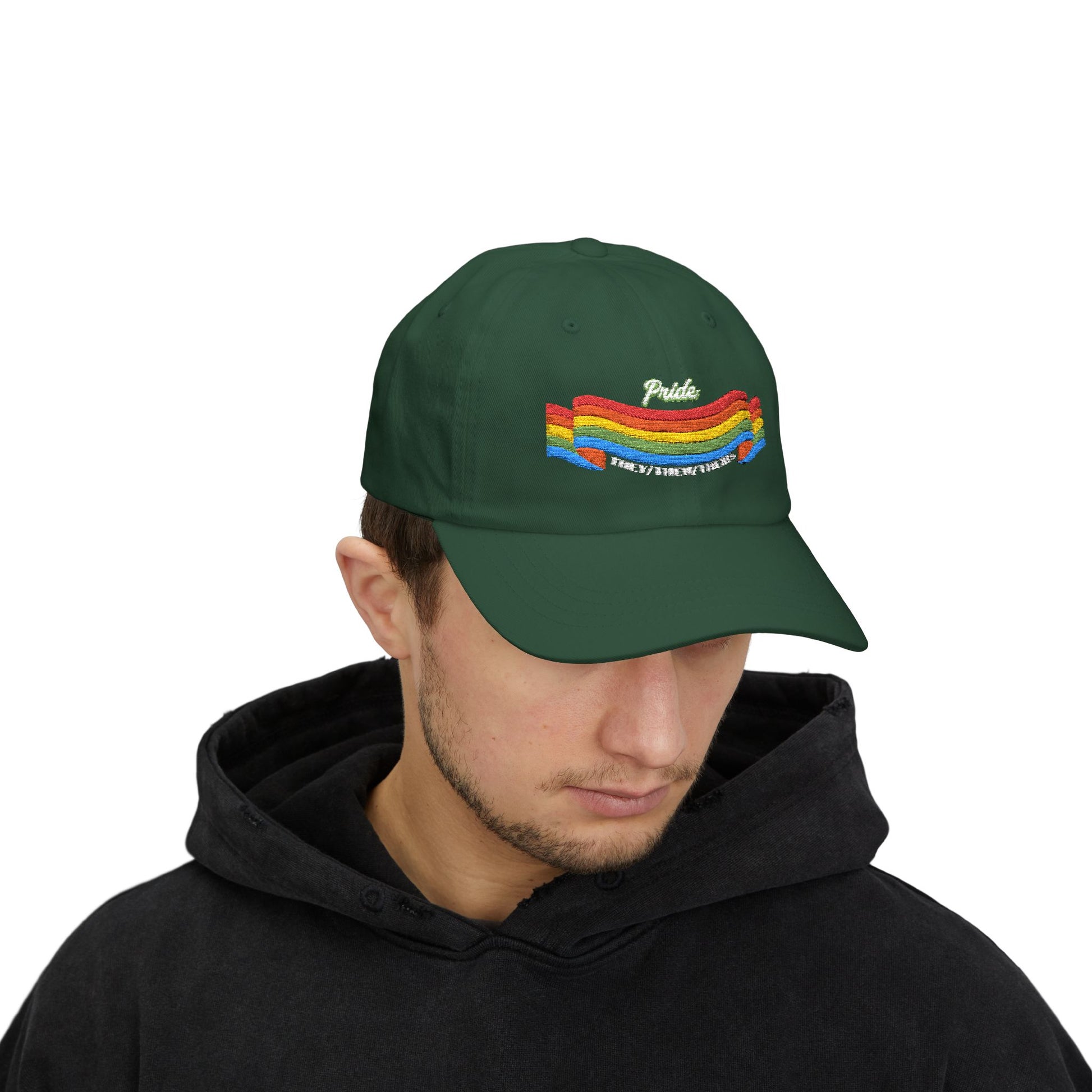 A person with short brown hair wears a green cap from the Worldwide PRIDE store, featuring a rainbow design and the embroidered pronouns they/them/theirs. The person is also wearing a black hooded sweatshirt and looking down.