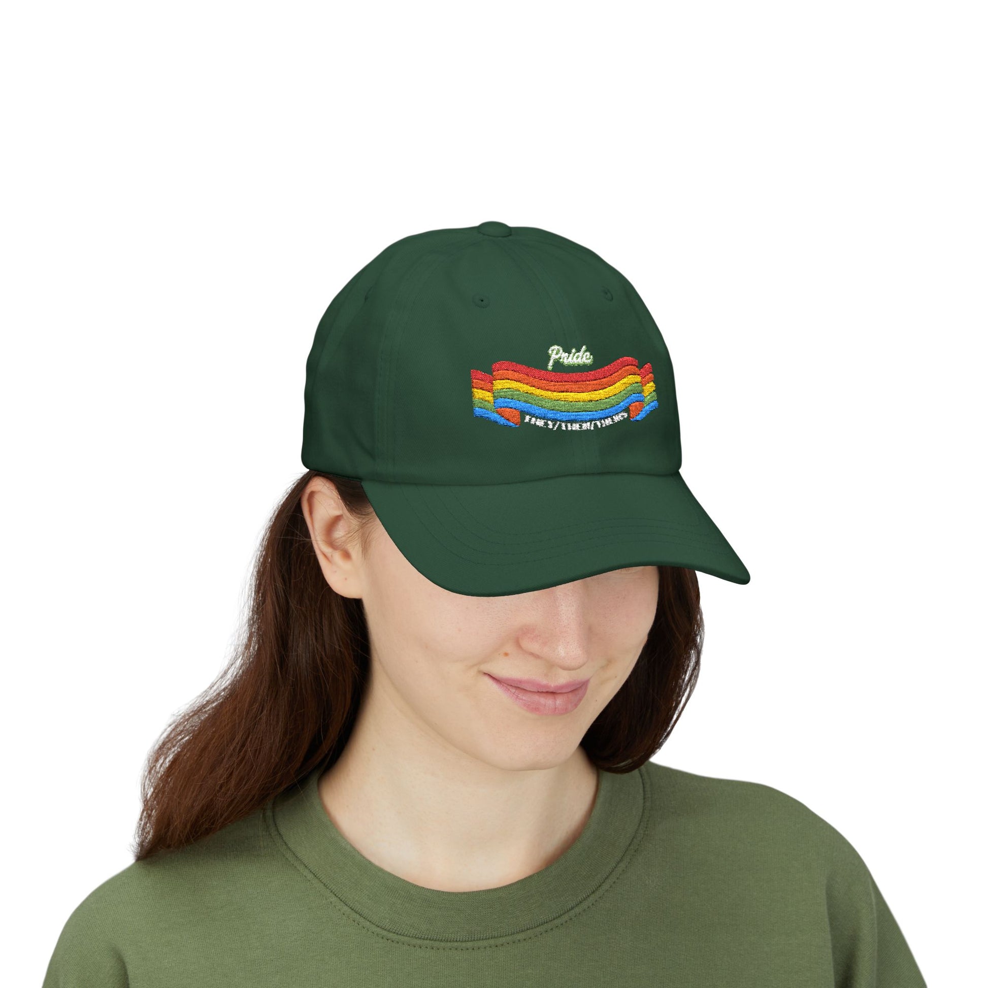 A person with long brown hair wears a green cap from the Worldwide PRIDE store, featuring a rainbow design and the embroidered pronouns they/them/theirs, along with an olive green sweatshirt. The person is smiling softly and looking down.