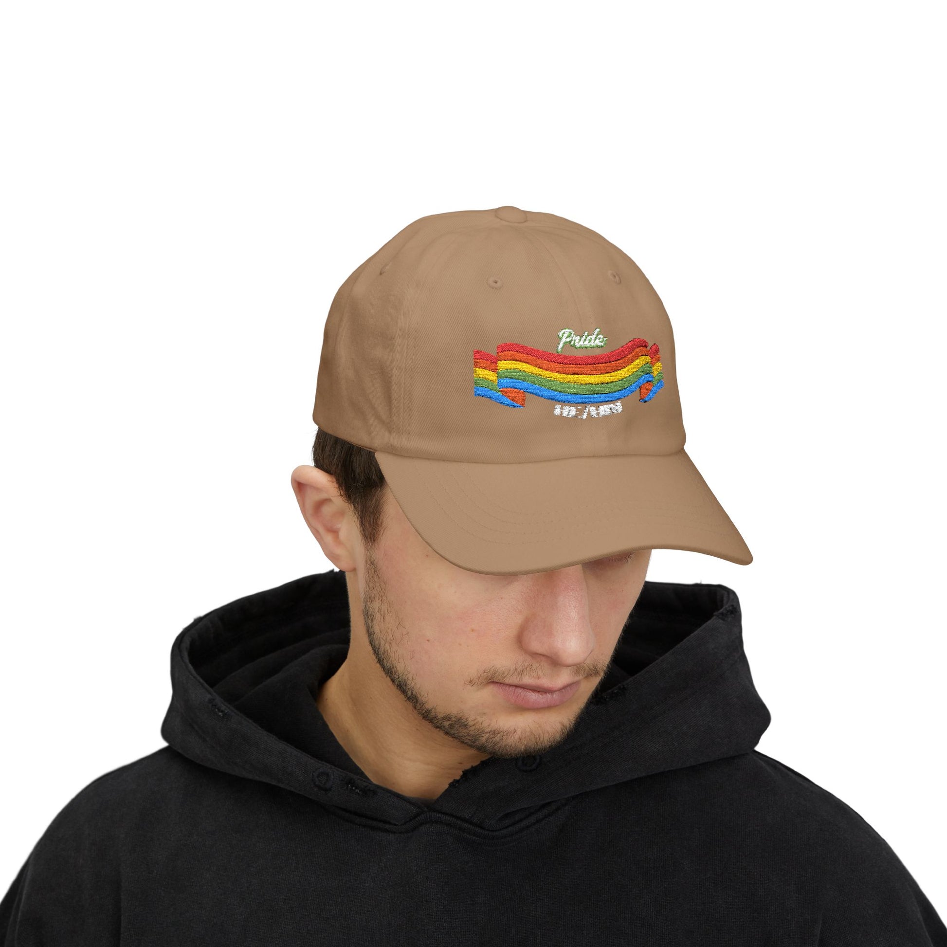 A person with short brown hair wears a khaki cap from the Worldwide PRIDE store, featuring a rainbow design and the embroidered pronouns he/him. The person is also wearing a black hooded sweatshirt and looking down.