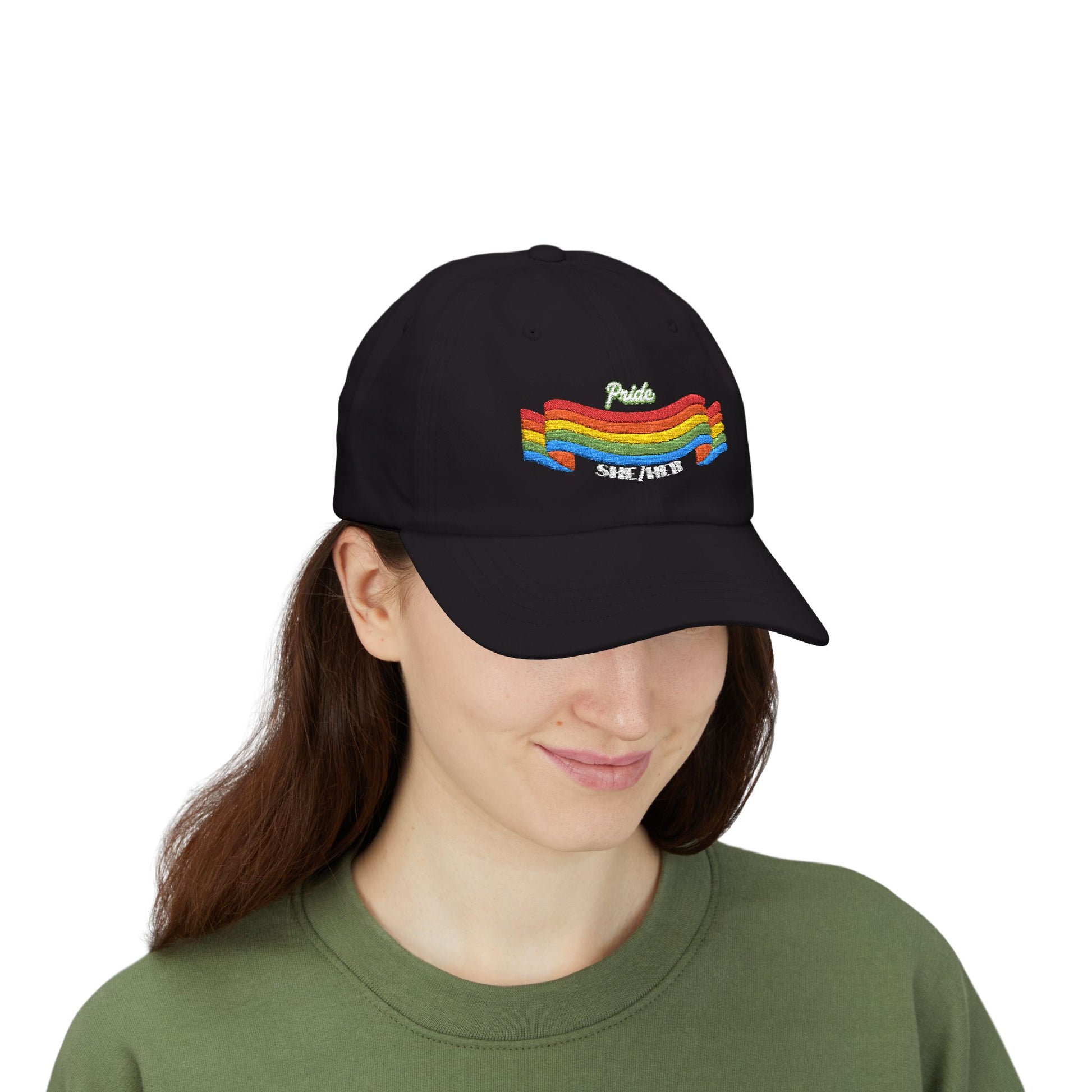 A person with long brown hair wears a black cap from the Worldwide PRIDE store, featuring a rainbow design and the embroidered pronouns she/her, along with an olive green sweatshirt. The person is smiling softly and looking down.