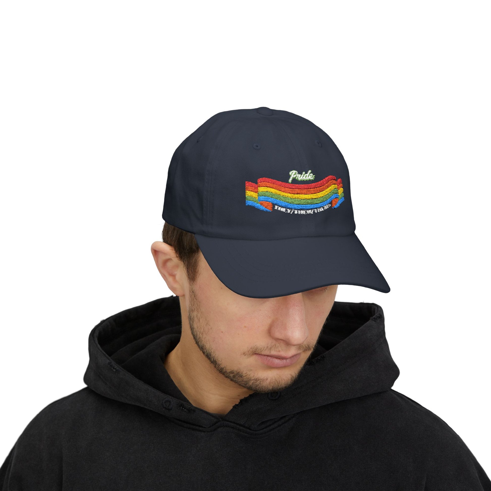 A person with short brown hair wears a navy blue cap from the Worldwide PRIDE store, featuring a rainbow design and the embroidered pronouns they/them/theirs. The person is also wearing a black hooded sweatshirt and looking down.