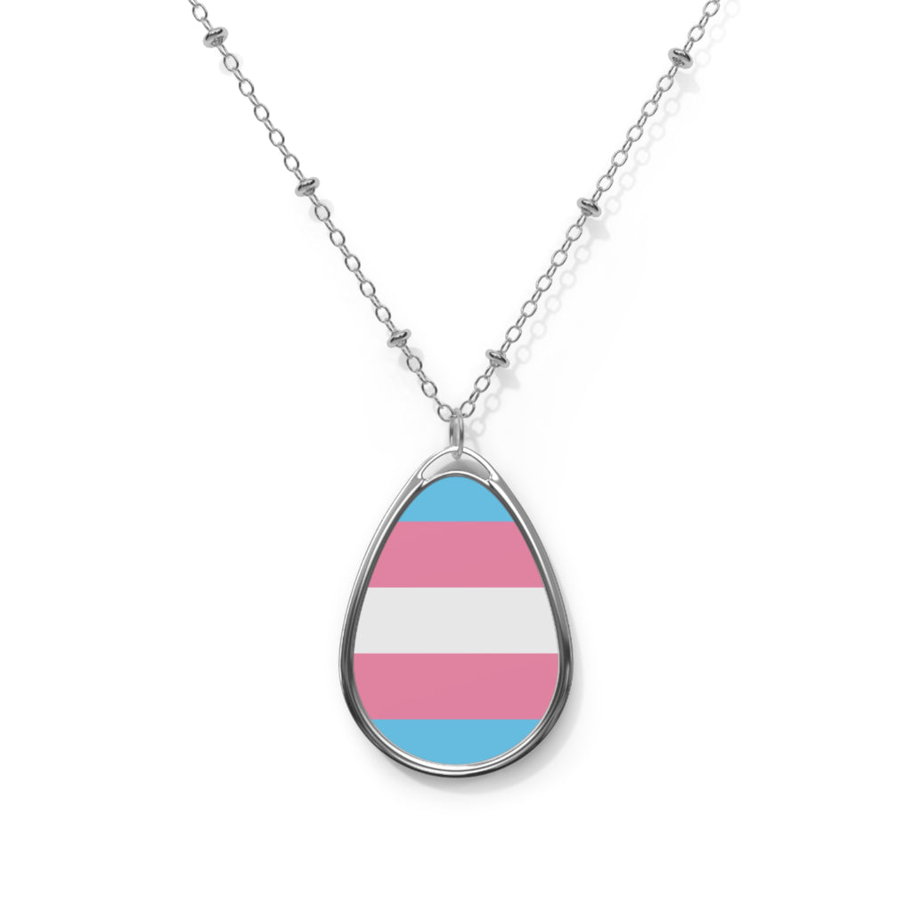 Close up of a teardrop shaped necklace proudly displaying the colors and design of the transsexual flag. The necklace hangs on a glass box. The carolina blue, pink and white colors symbolize the struggle and plight of the Trans community.