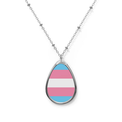 Close up of a teardrop shaped necklace proudly displaying the colors and design of the transsexual flag. The necklace hangs on a glass box. The carolina blue, pink and white colors symbolize the struggle and plight of the Trans community.