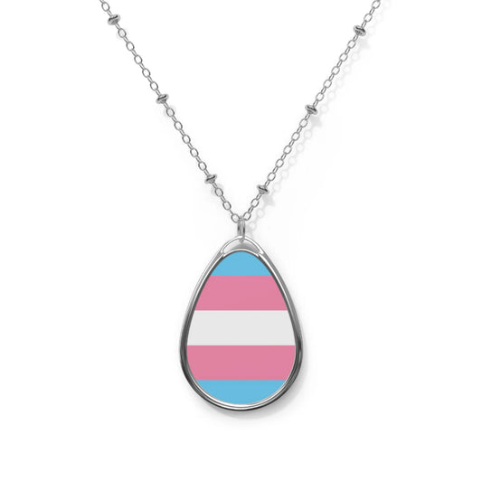 Close up of a teardrop shaped necklace proudly displaying the colors and design of the transsexual flag. The necklace hangs on a glass box. The carolina blue, pink and white colors symbolize the struggle and plight of the Trans community.