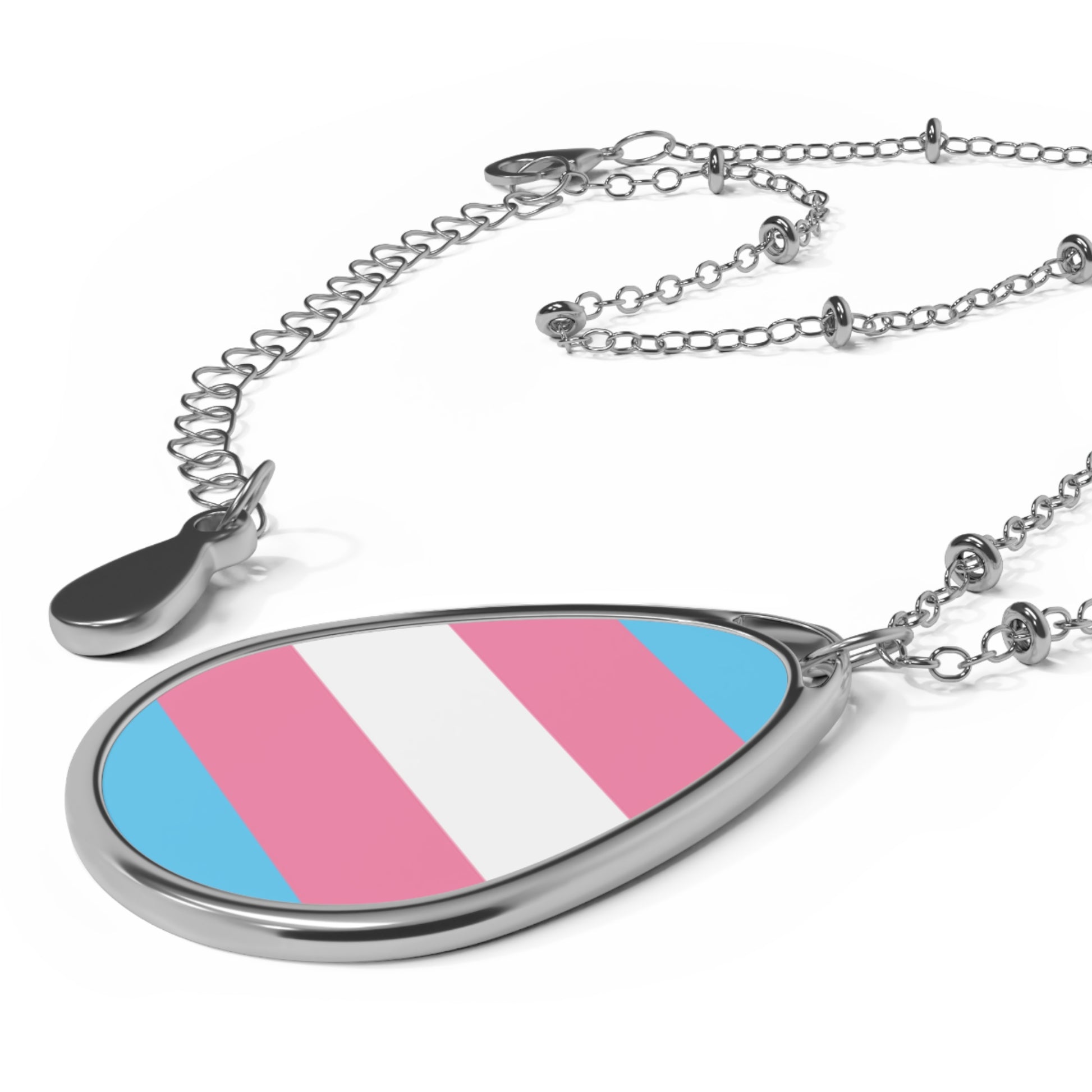Close up of a teardrop-shaped necklace proudly displaying the colors and design of the transsexual flag. The necklace sits on a white table. The carolina blue, pink and white colors symbolize the struggle and plight of the Trans community.