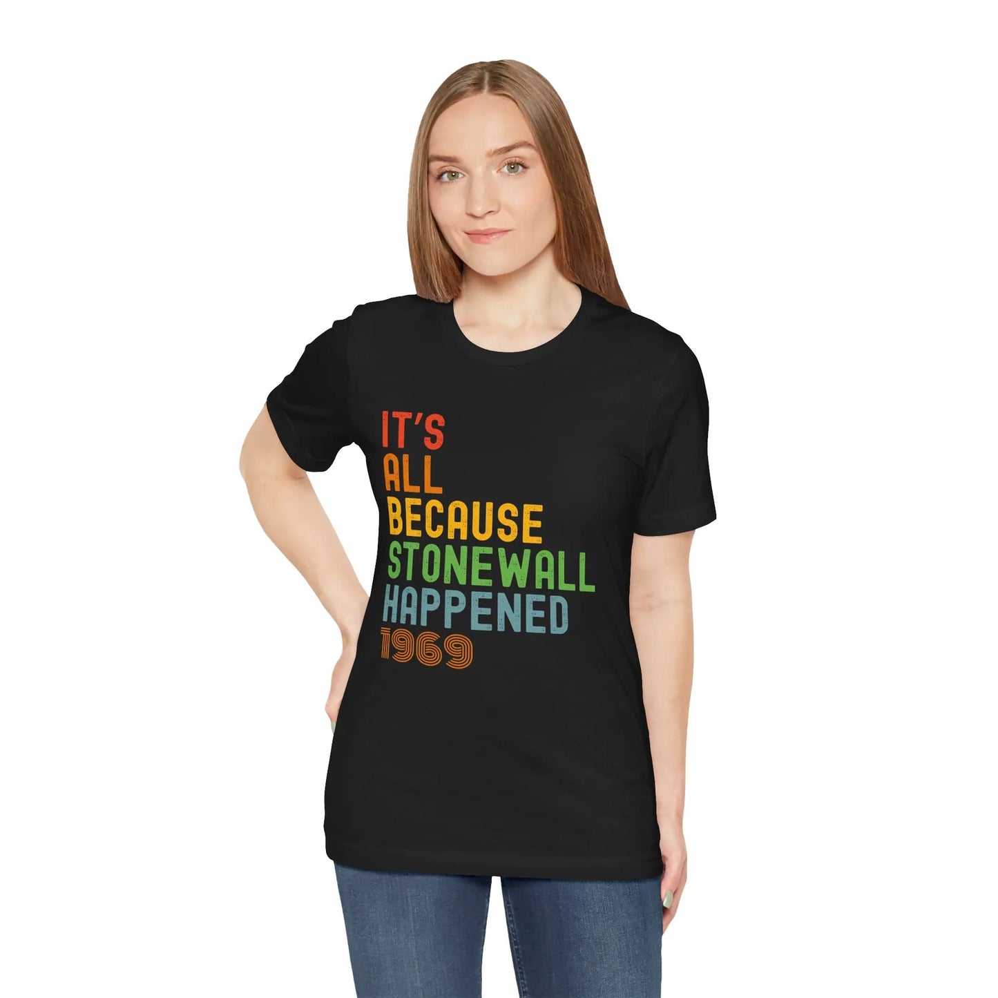 A person wearing a black unisex short-sleeve t-shirt with the text 'IT'S ALL BECAUSE STONEWALL HAPPENED' printed in bold, multicolored letters.