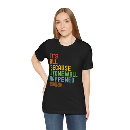 A person wearing a black unisex short-sleeve t-shirt with the text 'IT'S ALL BECAUSE STONEWALL HAPPENED' printed in bold, multicolored letters.