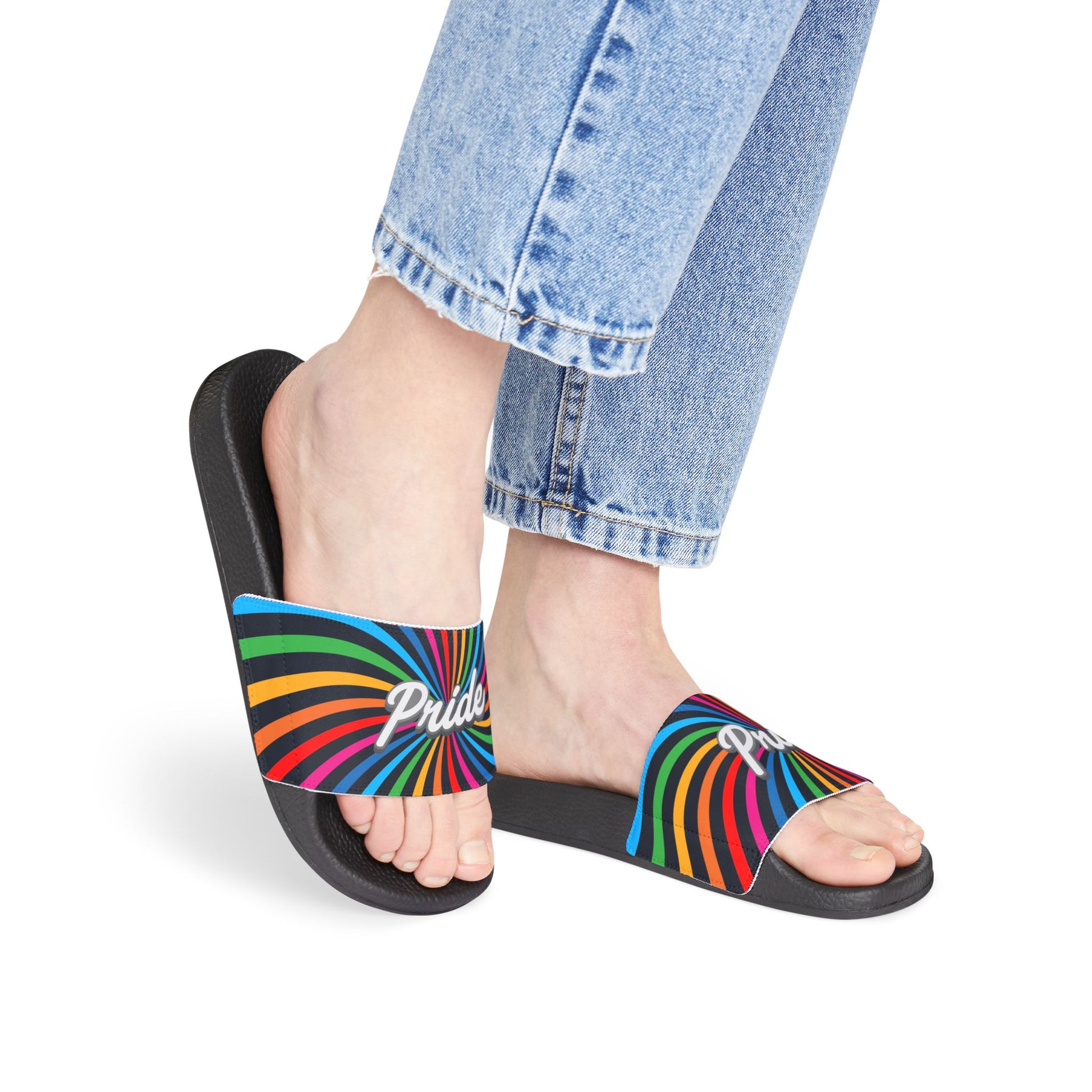 A person wearing light blue jeans stands donning black slides with rainbow stripes and the word Pride on them, celebrating Worldwide Pride. The background is plain white, highlighting their support for the LGBTQ community.