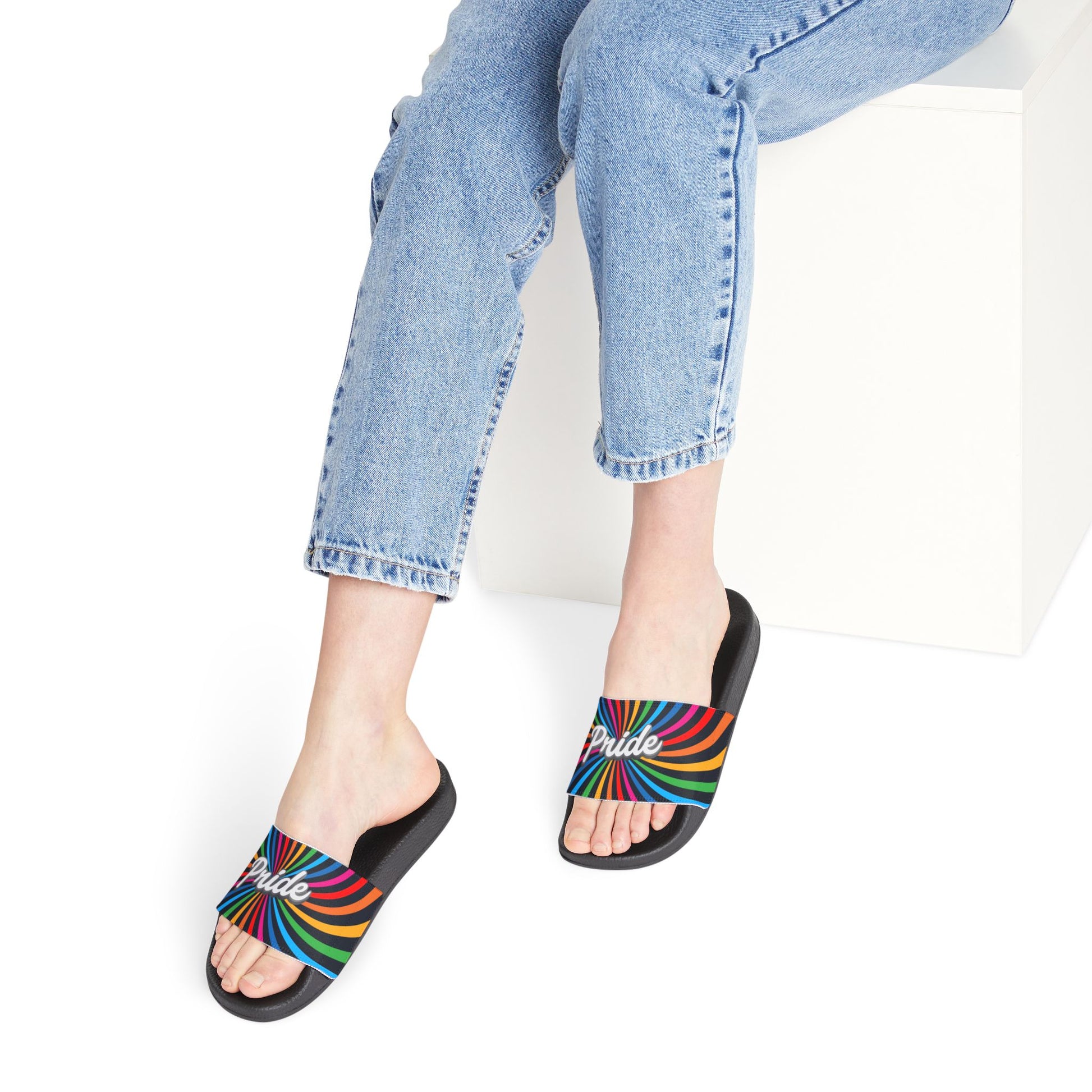 A person wearing light blue jeans sits on a white block, donning black slides with rainbow stripes and the word Pride on them, celebrating Worldwide Pride. The background is plain white, highlighting their support for the LGBTQ community.