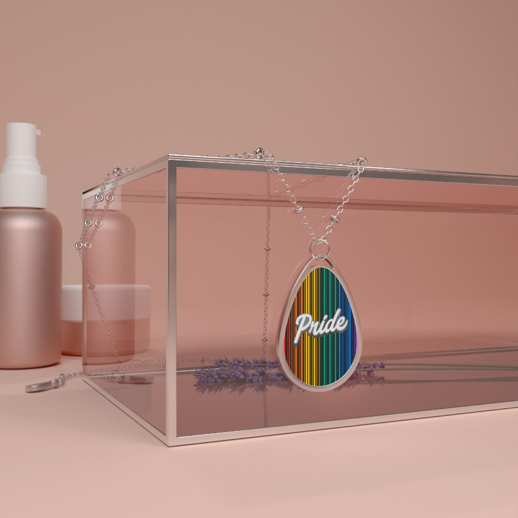 A silver chain necklace showcases a teardrop-shaped pendant adorned with a rainbow-striped pattern and the word PRIDE in elegant white cursive. The necklace hangs over a glass box. The close-up captures the vibrant colors and shiny metal beautifully.