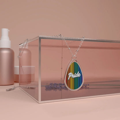 A silver chain necklace showcases a teardrop-shaped pendant adorned with a rainbow-striped pattern and the word PRIDE in elegant white cursive. The necklace hangs over a glass box. The close-up captures the vibrant colors and shiny metal beautifully.