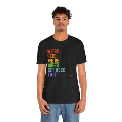 A person wearing a black unisex crew neck t-shirt with the text 'WE'RE HERE WE'RE QUEER GET USED TO IT' printed in multicolored letters.