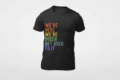 A black unisex crew neck t-shirt with the text 'WE'RE HERE WE'RE QUEER GET USED TO IT' printed in multicolored letters.