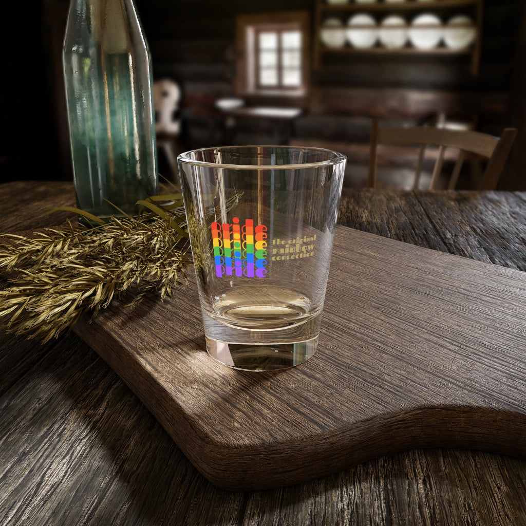 A shot glass features pride in a rainbow gradient on the left, celebrating the LGBTQ community. On the right, it reads The original rainbow connection in gold text. The glass sits on a wooden cutting board with a empty bottle in the background.