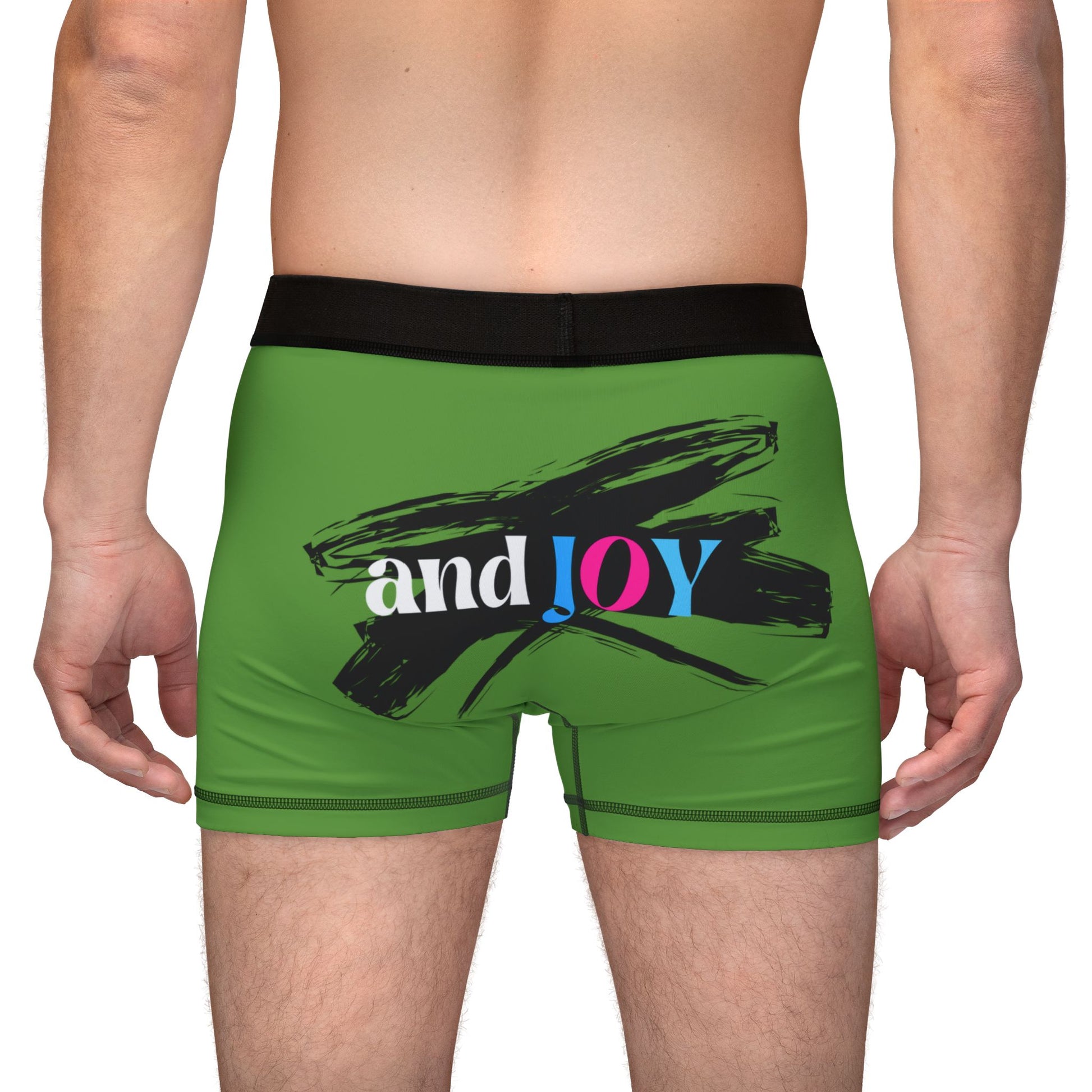 Rear view of a person wearing green boxer shorts with a black waistband. The design boasts a black brushstroke and the words and JOY in white, pink, and blue on the back. Perfectly encapsulating Worldwide PRIDE, they stand confidently against a plain background.