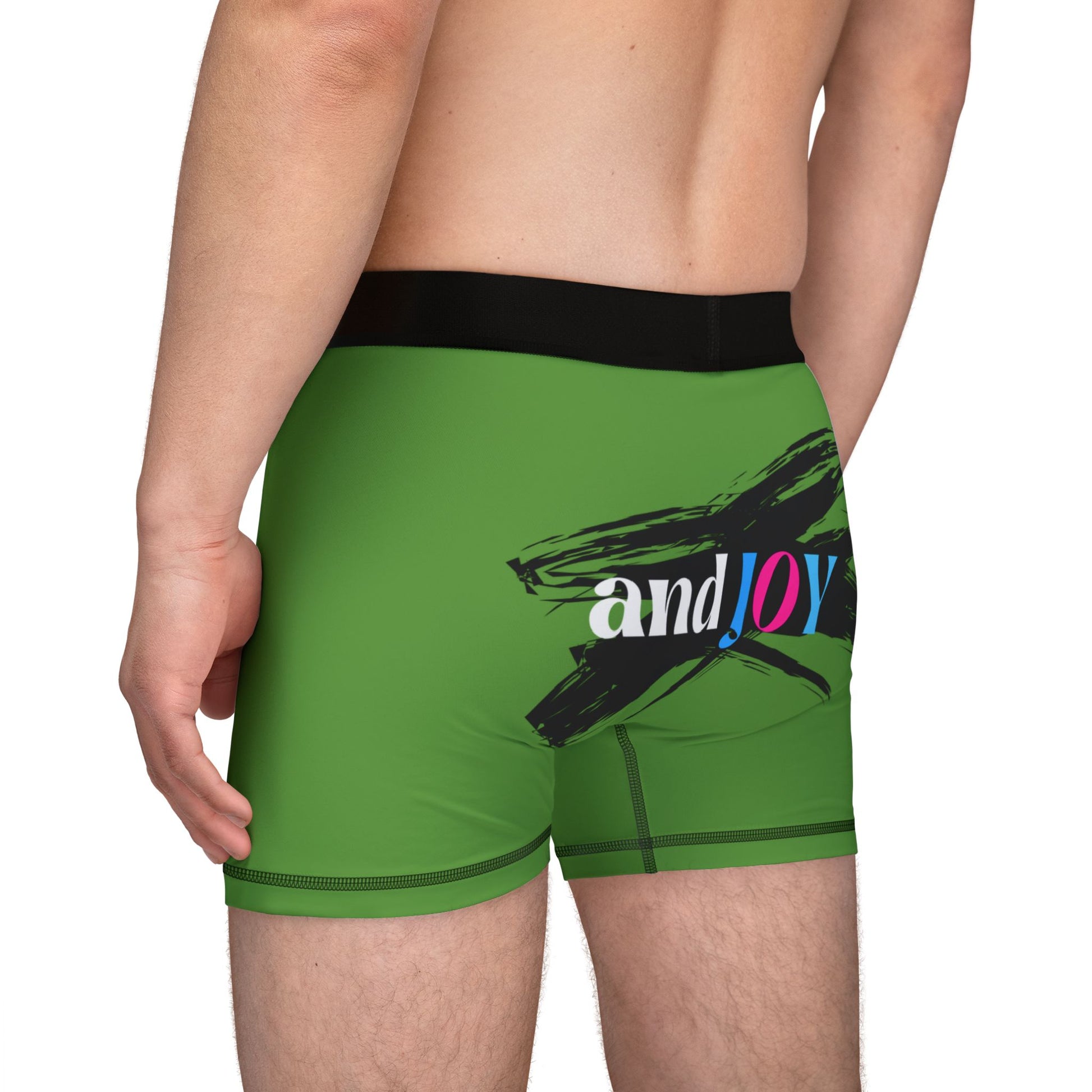 Rear view of a person wearing green boxer shorts with a black waistband. The design boasts a black brushstroke and the words and JOY in white, pink, and blue on the back. Perfectly encapsulating Worldwide PRIDE, they stand confidently against a plain background.
