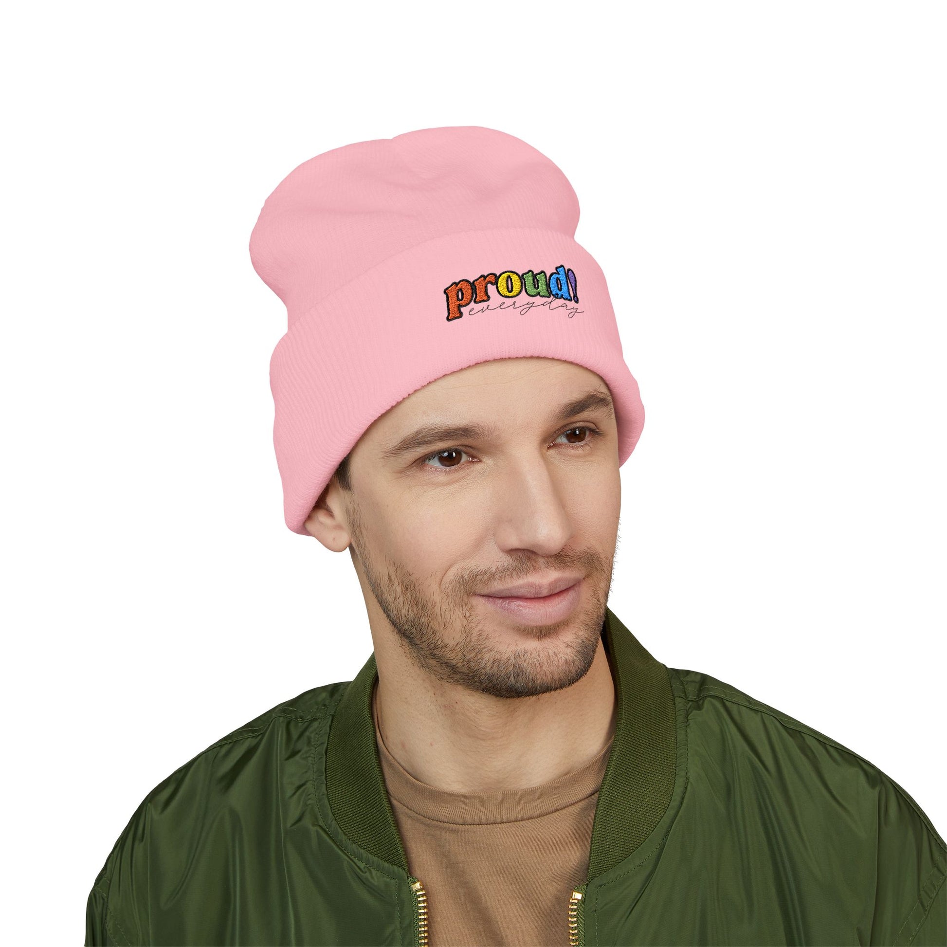 A person wearing a light pink beanie with “proud everyday" embroidered in rainbow colors and a green jacket, looking slightly to the side with a neutral expression. Perfect for the LGBTQ community from the Worldwide PRIDE store.