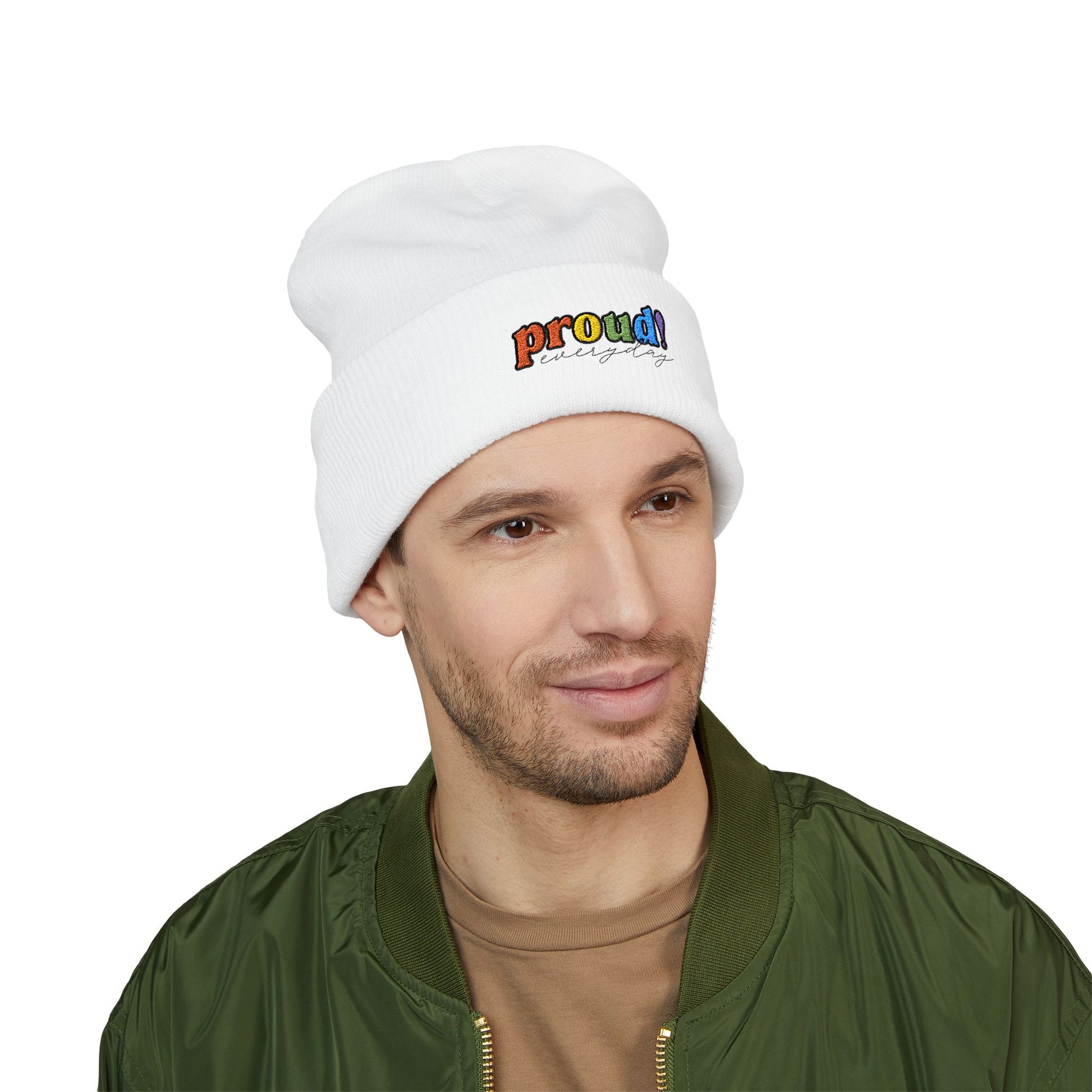 A person wearing a white beanie with “proud everyday" embroidered in rainbow colors and a green jacket, looking slightly to the side with a neutral expression. Perfect for the LGBTQ community from the Worldwide PRIDE store.