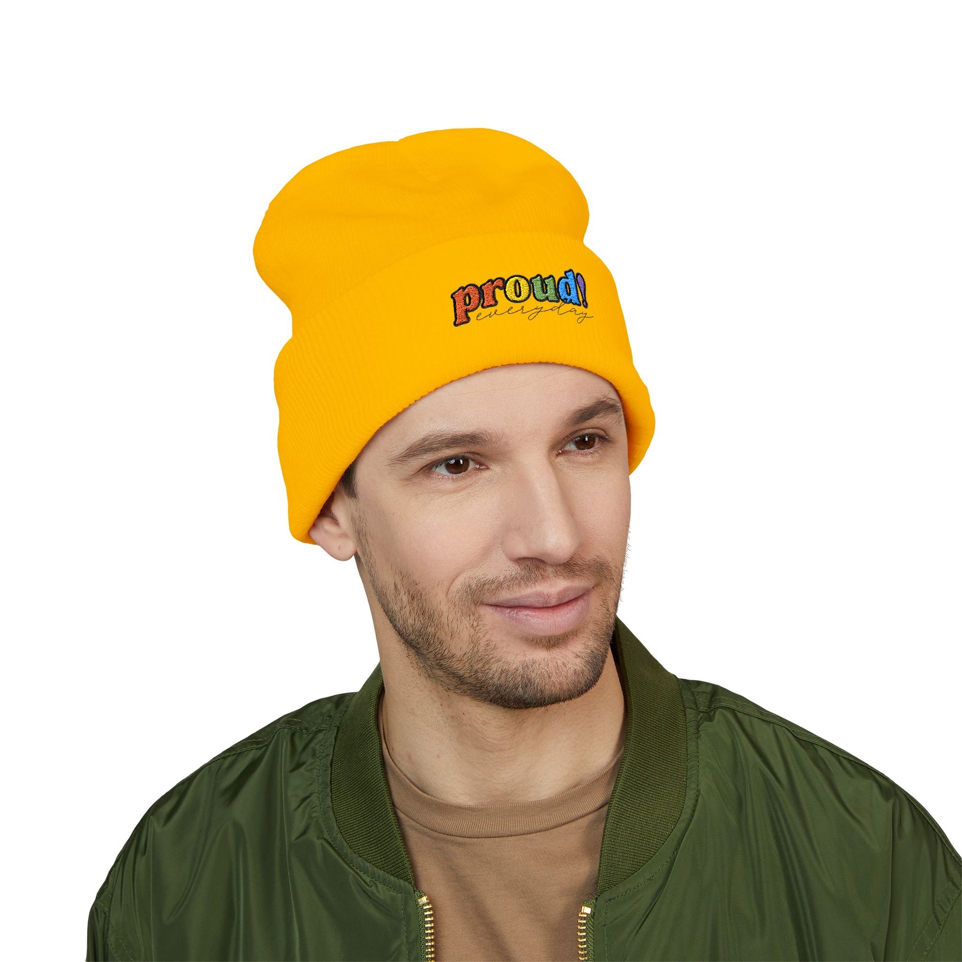 A person wearing a yellow beanie with “proud everyday" embroidered in rainbow colors and a green jacket, looking slightly to the side with a neutral expression. Perfect for the LGBTQ community from the Worldwide PRIDE store.