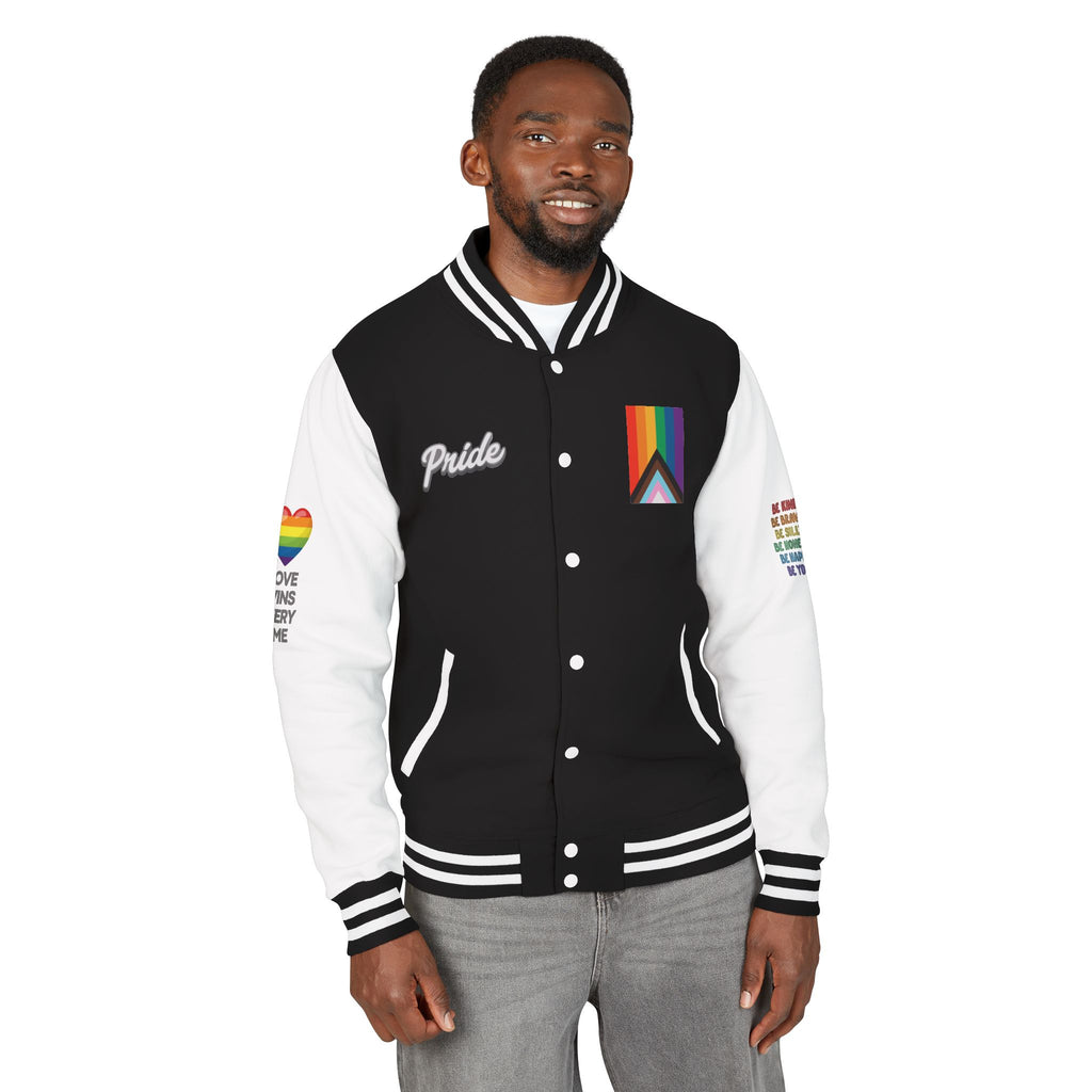 A person stands smiling, wearing a black and white letterman jacket from the Worldwide PRIDE store, adorned with a rainbow heart above the words "love wins every time" on the right sleeve, and the progress PRIDE flag, and the word PRIDE on the chest. The left sleeve is obstructed but reads: Be kind, be brave, be silly, be honest, be happy, be you.