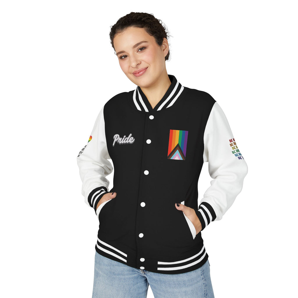A person stands smiling, wearing a black and white letterman jacket from the Worldwide PRIDE store, adorned with a rainbow heart above the words "love wins every time" on the right sleeve, and the progress PRIDE flag, and the word PRIDE on the chest. The left sleeve is obstructed but reads: Be kind, be brave, be silly, be honest, be happy, be you.