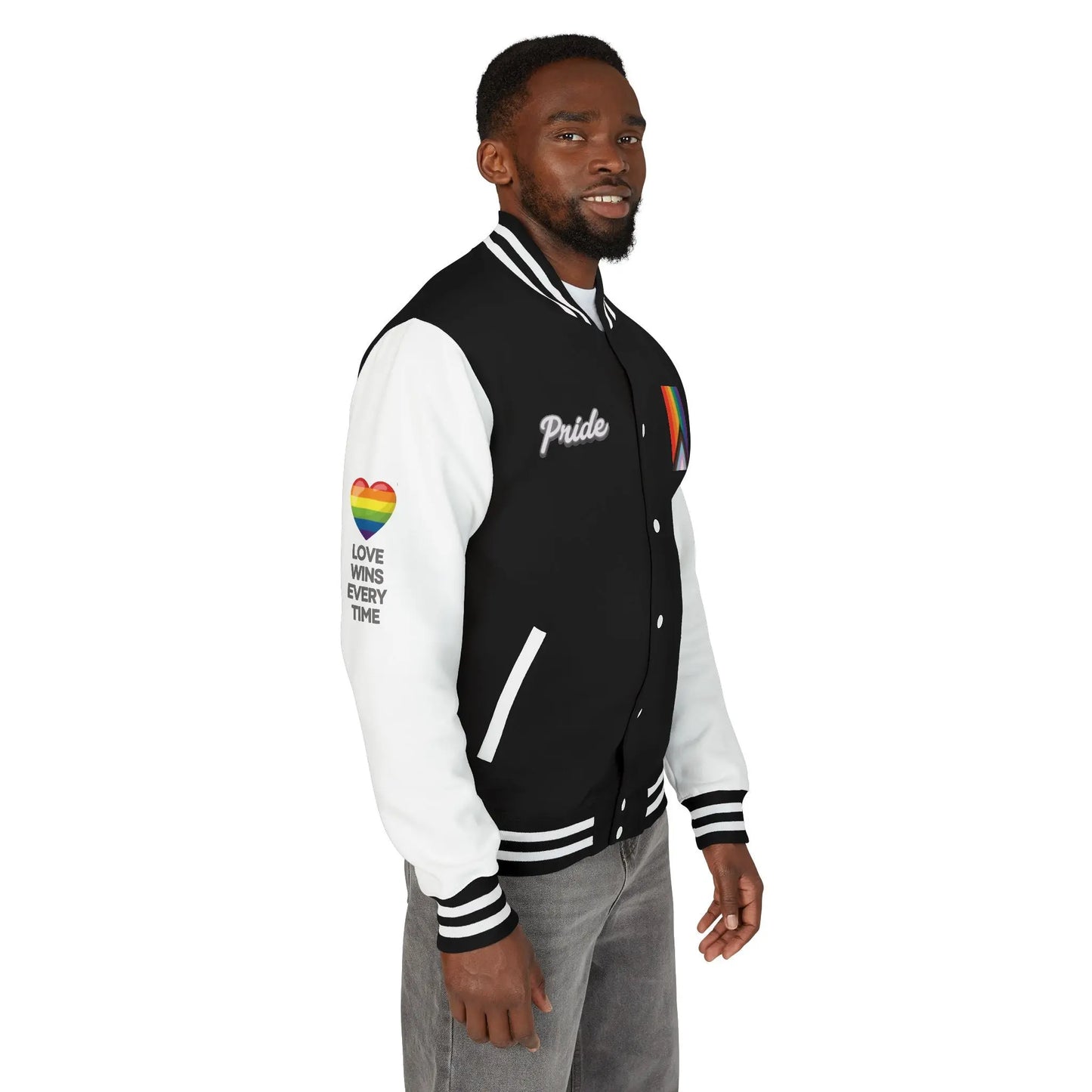 Right side view of a person stands smiling, wearing a black and white letterman jacket from the Worldwide PRIDE store, adorned with a rainbow heart above the words "love wins every time" on the right sleeve, and the progress PRIDE flag, and the word PRIDE on the chest. The left sleeve is obstructed but reads: Be kind, be brave, be silly, be honest, be happy, be you.