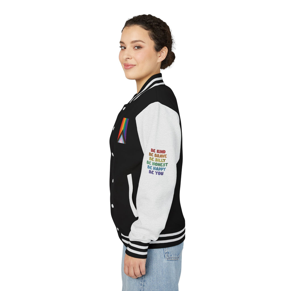 Left side view of a person who stands smiling, wearing a black and white letterman jacket from the Worldwide PRIDE store, adorned with a rainbow heart above the words "love wins every time" on the right sleeve, and the progress PRIDE flag, and the word PRIDE on the chest. The left sleeve reads: Be kind, be brave, be silly, be honest, be happy, be you.