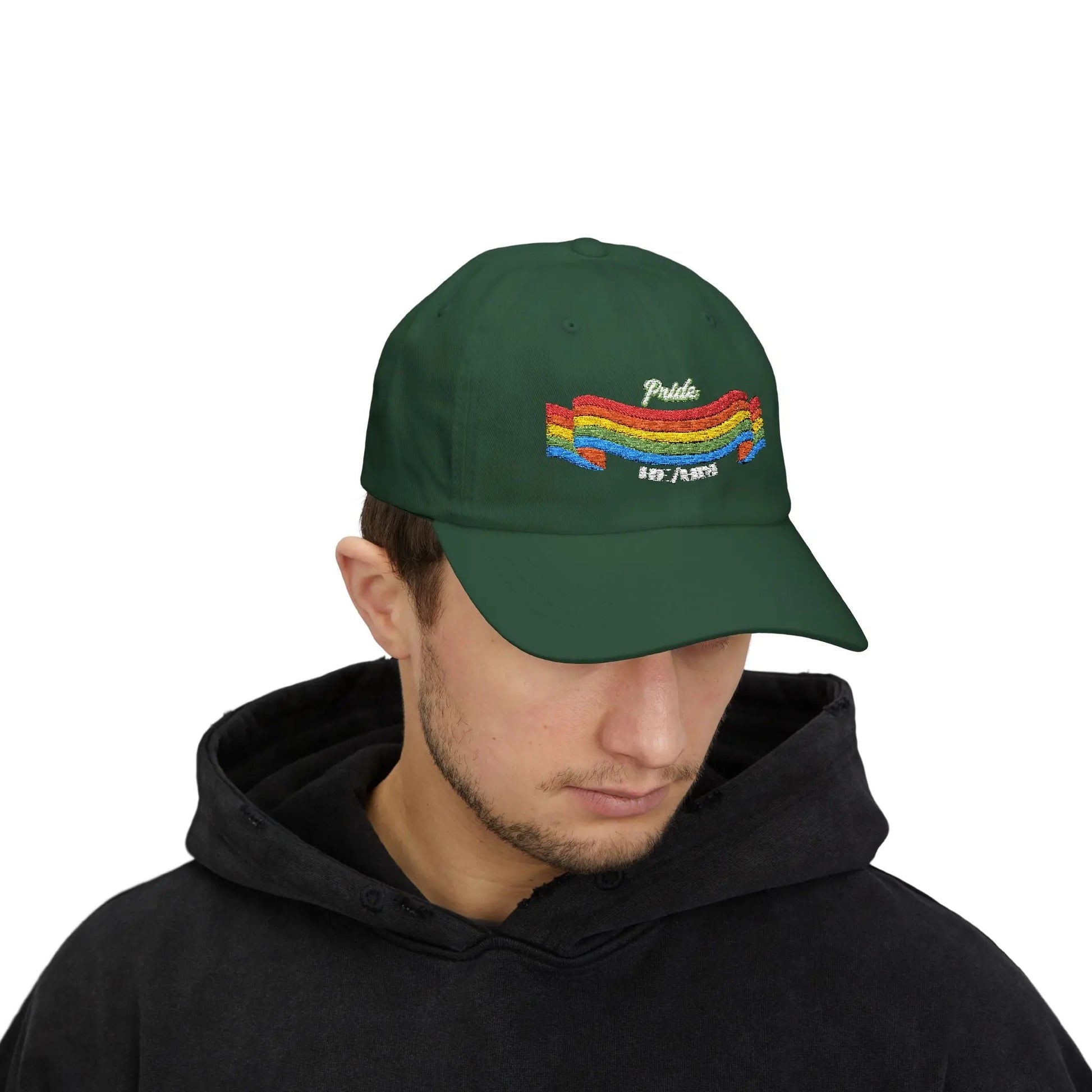A person with short brown hair wears a green cap from the Worldwide PRIDE store, featuring a rainbow design and the embroidered pronouns he/him.