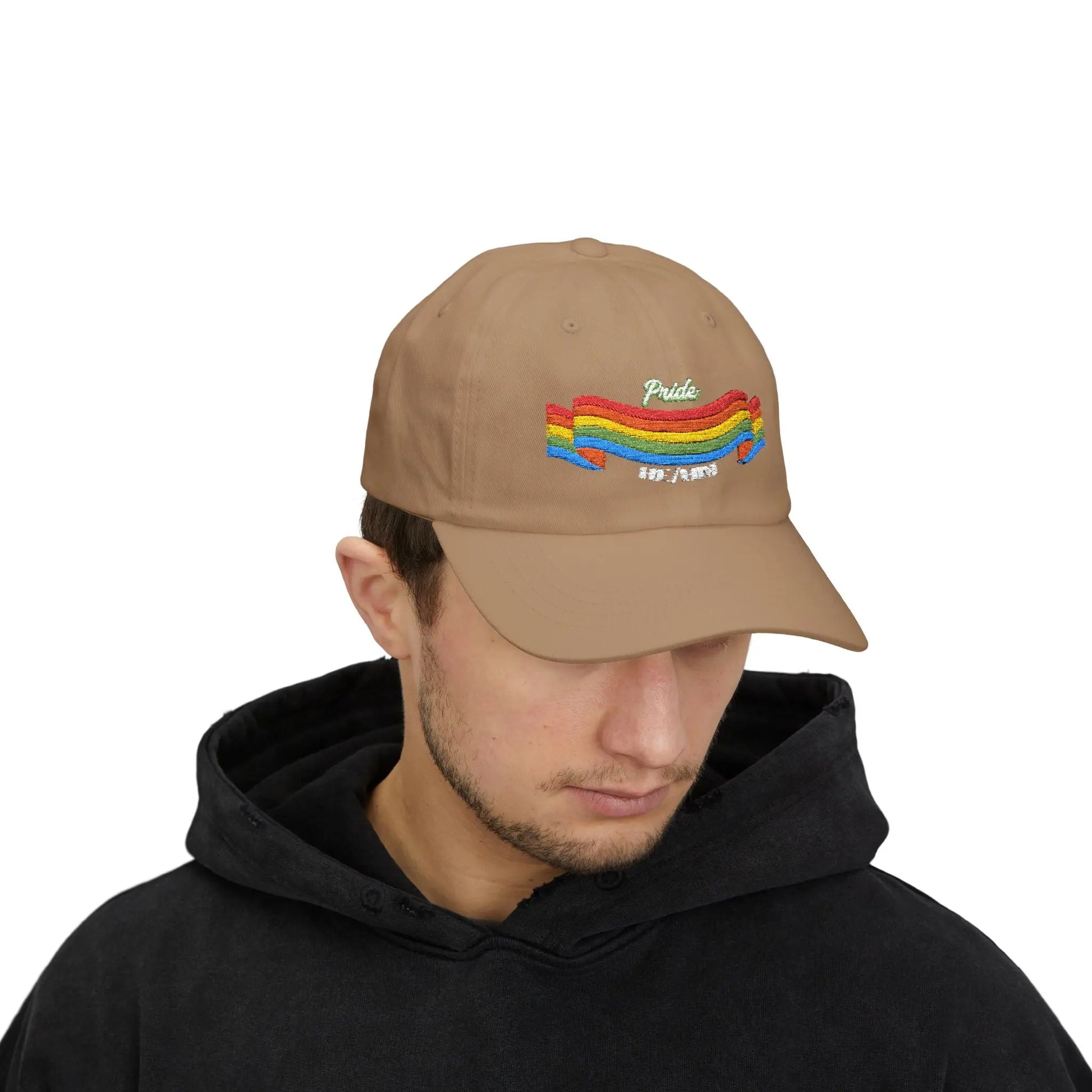 A person with short brown hair wears a khaki cap from the Worldwide PRIDE store, featuring a rainbow design and the embroidered pronouns he/him.