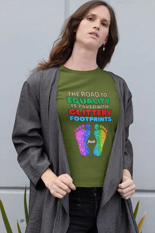 A person wearing an olive green unisex short-sleeve t-shirt with a graphic of multicolored glitter footprints and the text 'THE ROAD TO EQUALITY IS LITTERED WITH GLITTERY FOOTPRINTS'.