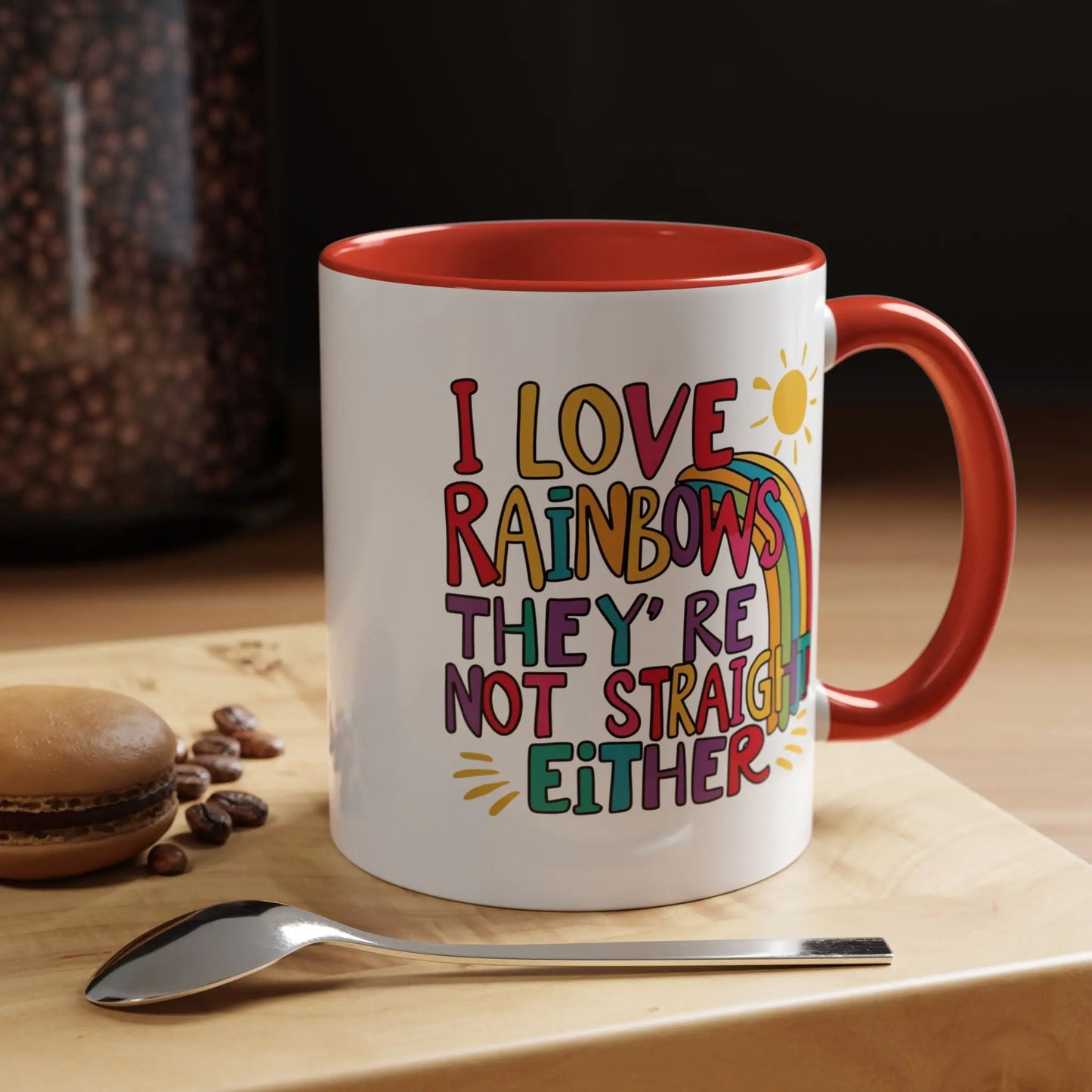 A white ceramic coffee mug with a red handle and interior, featuring the colorful text 'I LOVE RAINBOWS THEY'RE NOT STRAIGHT EITHER' along with a graphic of a rainbow and a sun.